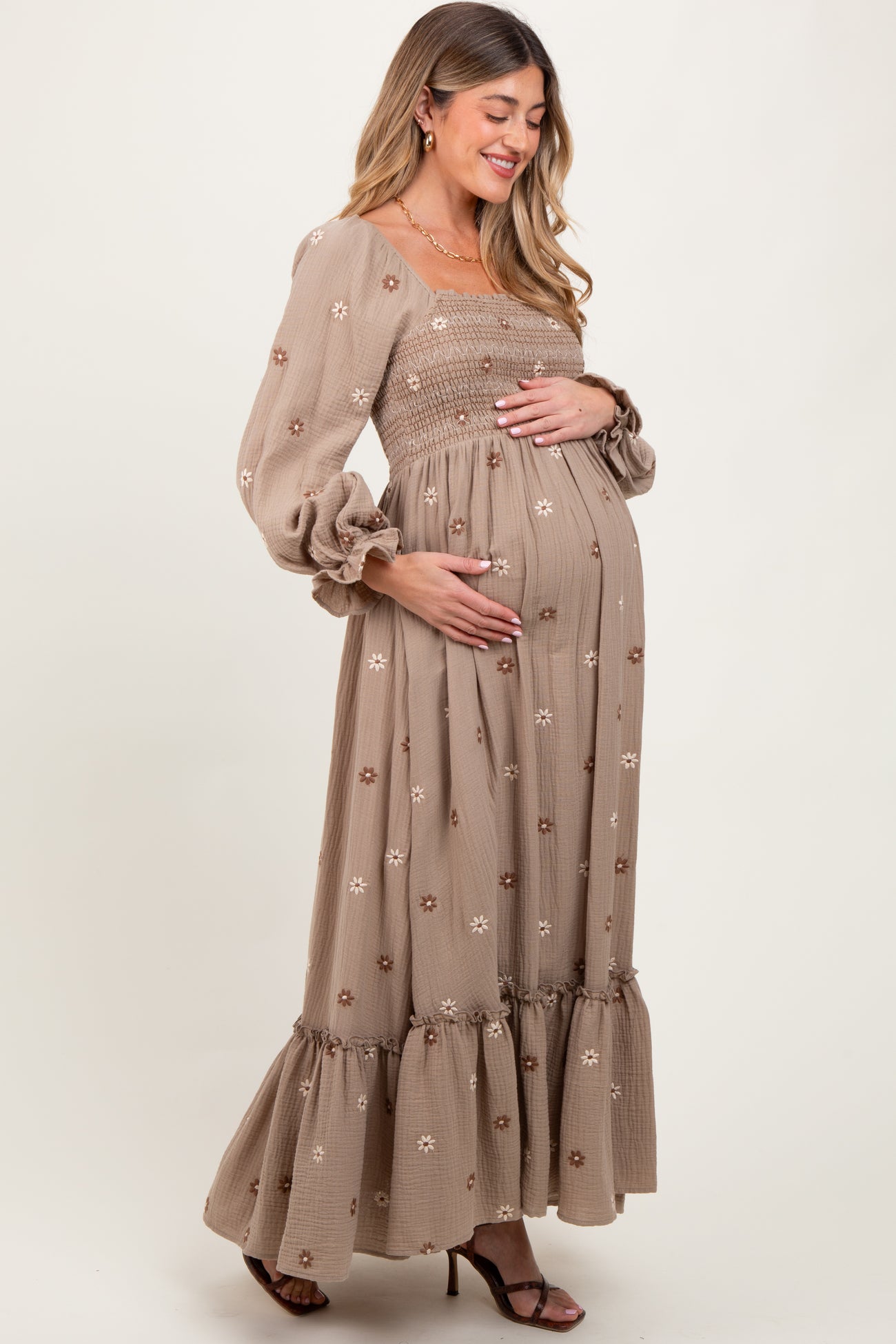 woman wearing a taupe long sleeve maternity dress with floral embroidery and ruffle hem, cradling her baby bump