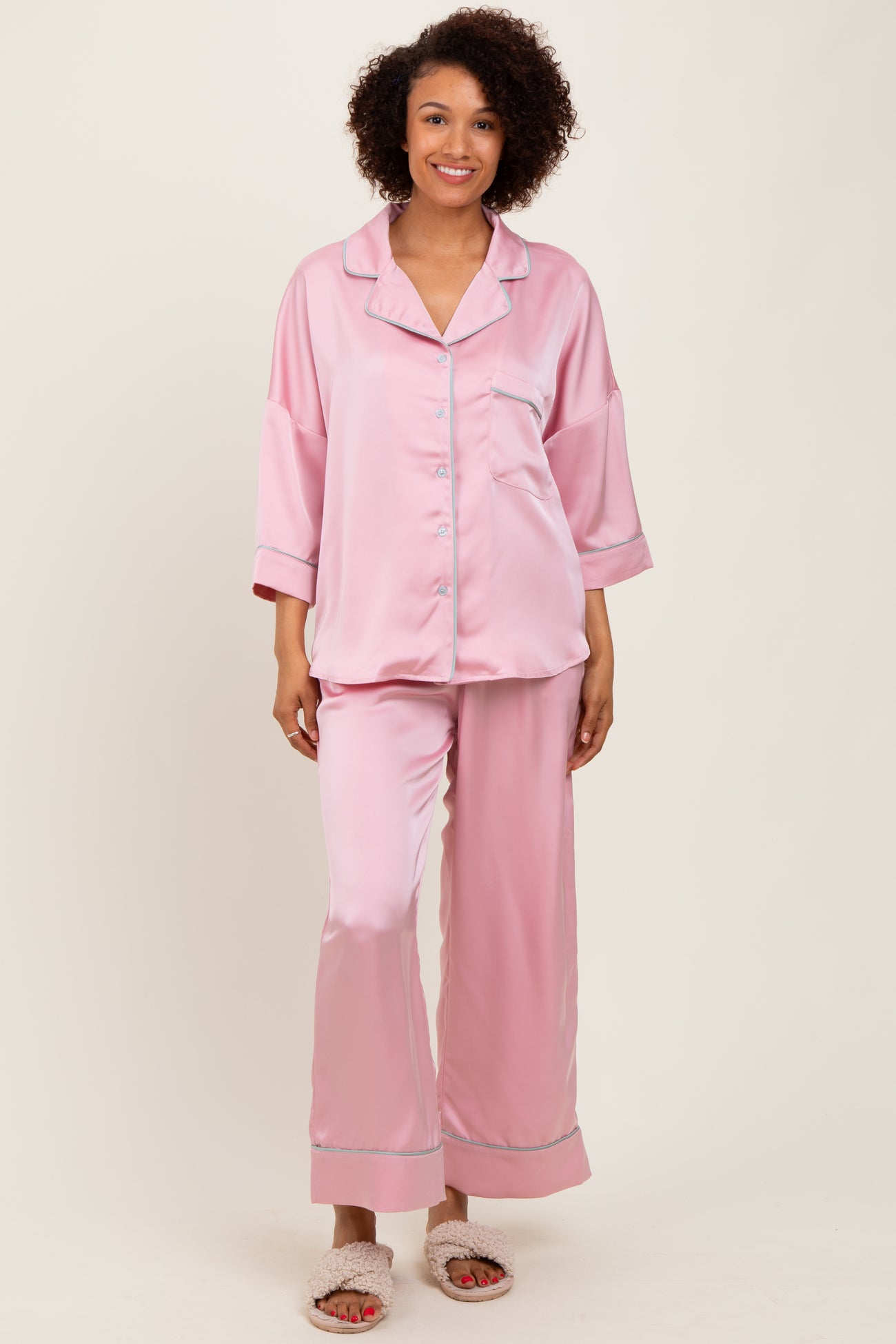 woman wearing a light pink satin pajama set with button-up top and wide-leg pants, standing and smiling