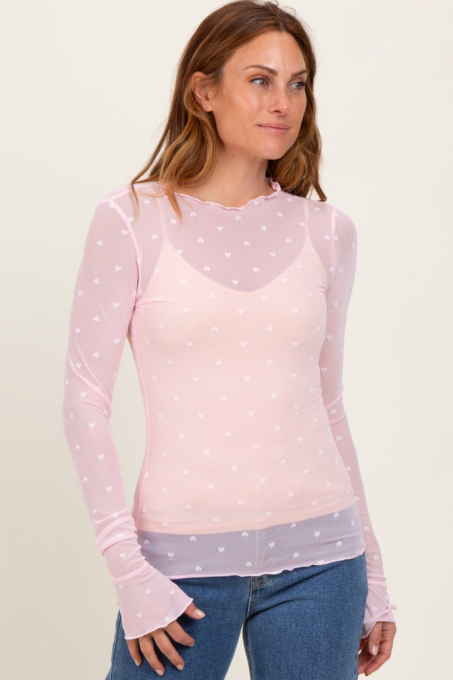 woman wearing a light pink long sleeve top with white heart print and ruffled neckline, styled with denim jeans