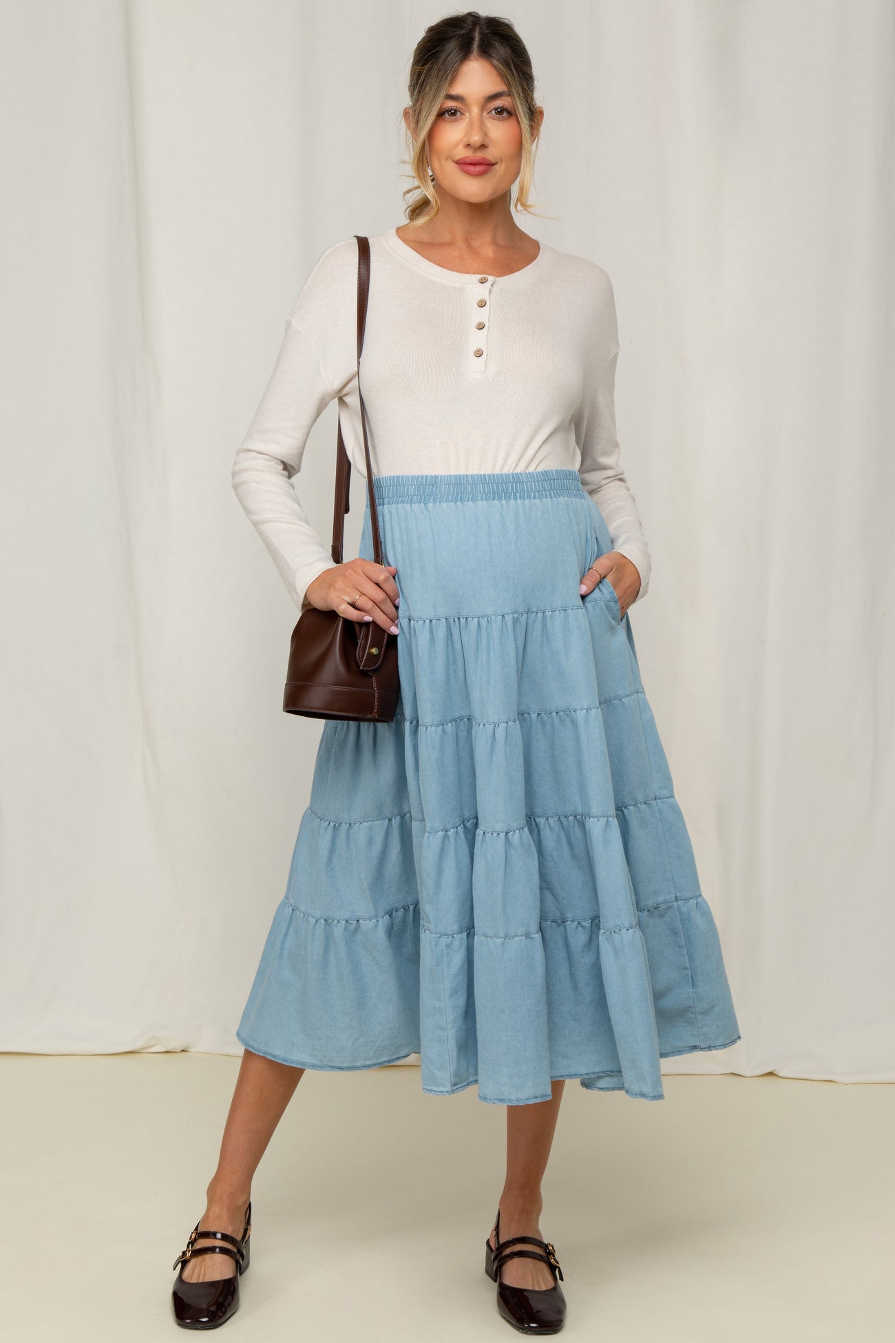 woman wearing a cream long sleeve henley top and a light blue tiered midi maternity skirt, holding a brown handbag