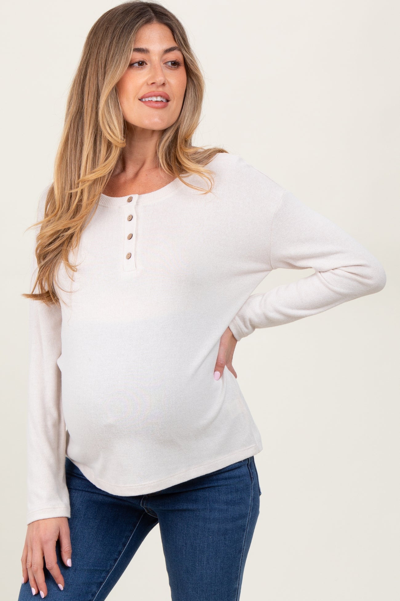 woman wearing a light beige maternity top with button details and a visible baby bump, paired with blue jeans