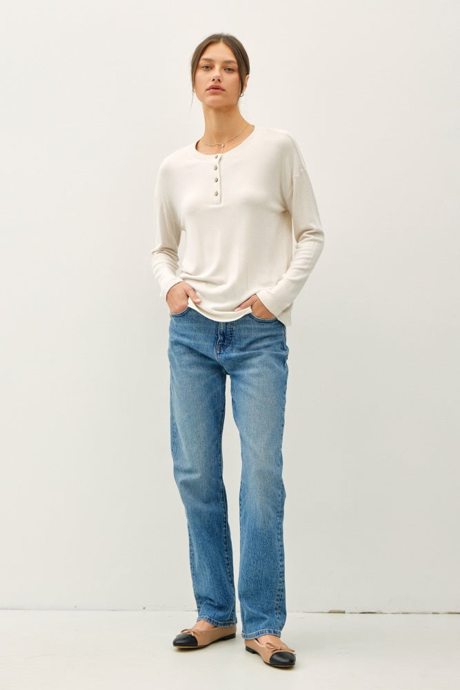 woman wearing a cream long sleeve henley top with buttons and blue straight-leg jeans, standing with hands in pockets