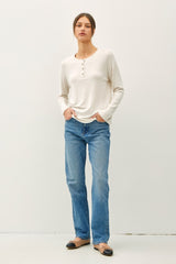 woman wearing a cream long sleeve henley top with buttons and blue straight-leg jeans, standing with hands in pockets