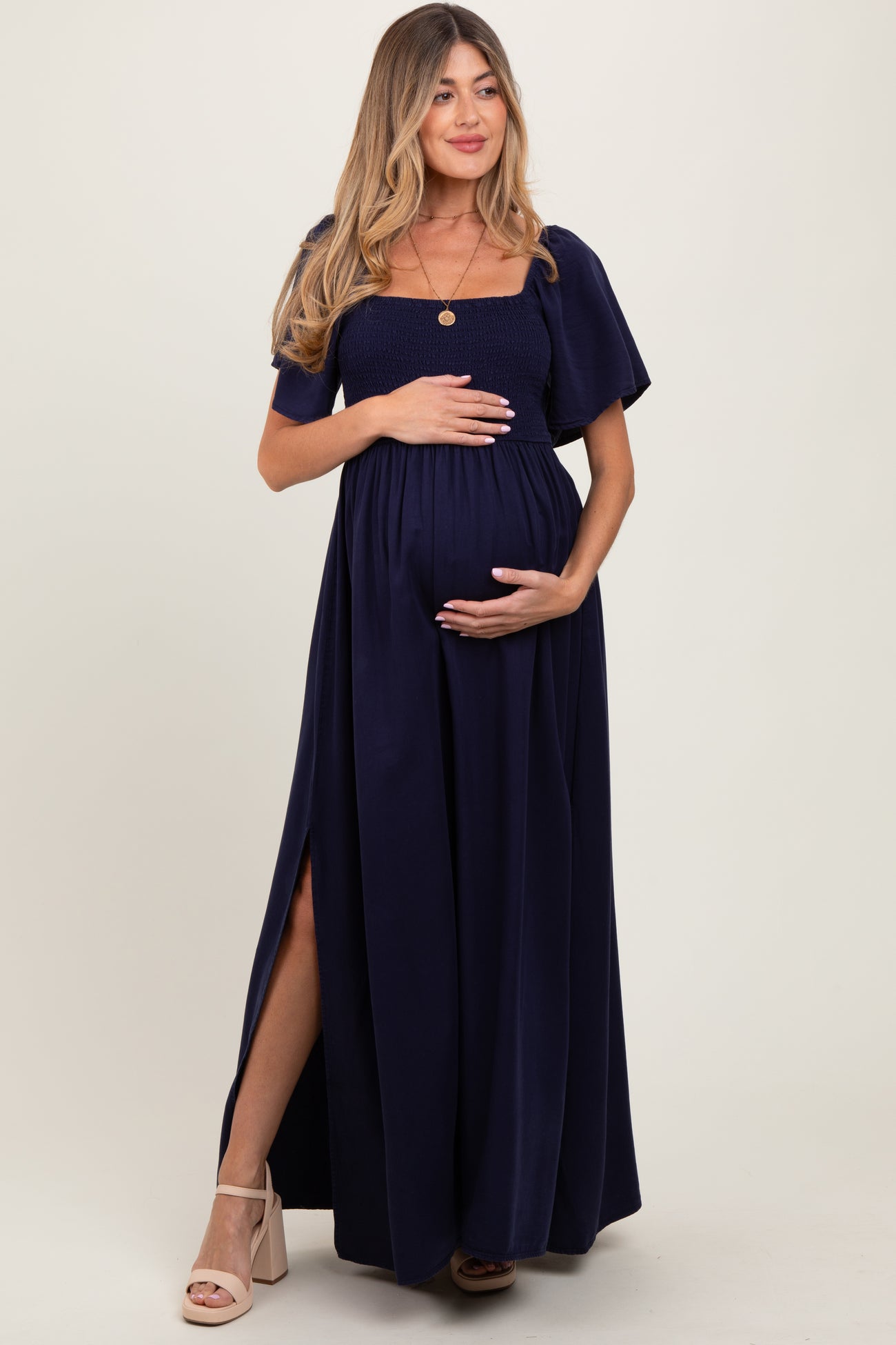 woman wearing a navy blue maternity maxi dress with a smocked bodice and a side slit, holding her baby bump