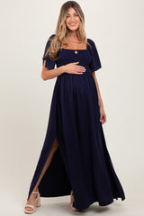 woman wearing a navy blue maternity maxi dress with a smocked bodice and a side slit, holding her baby bump
