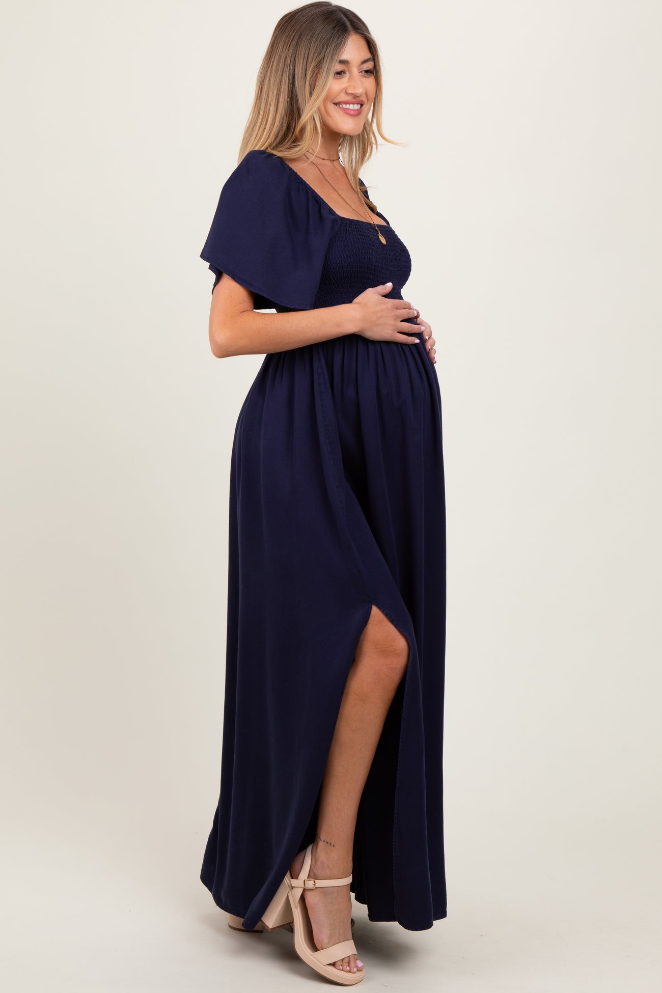 woman wearing a navy blue maternity maxi dress with a smocked bodice and side slit, holding her baby bump