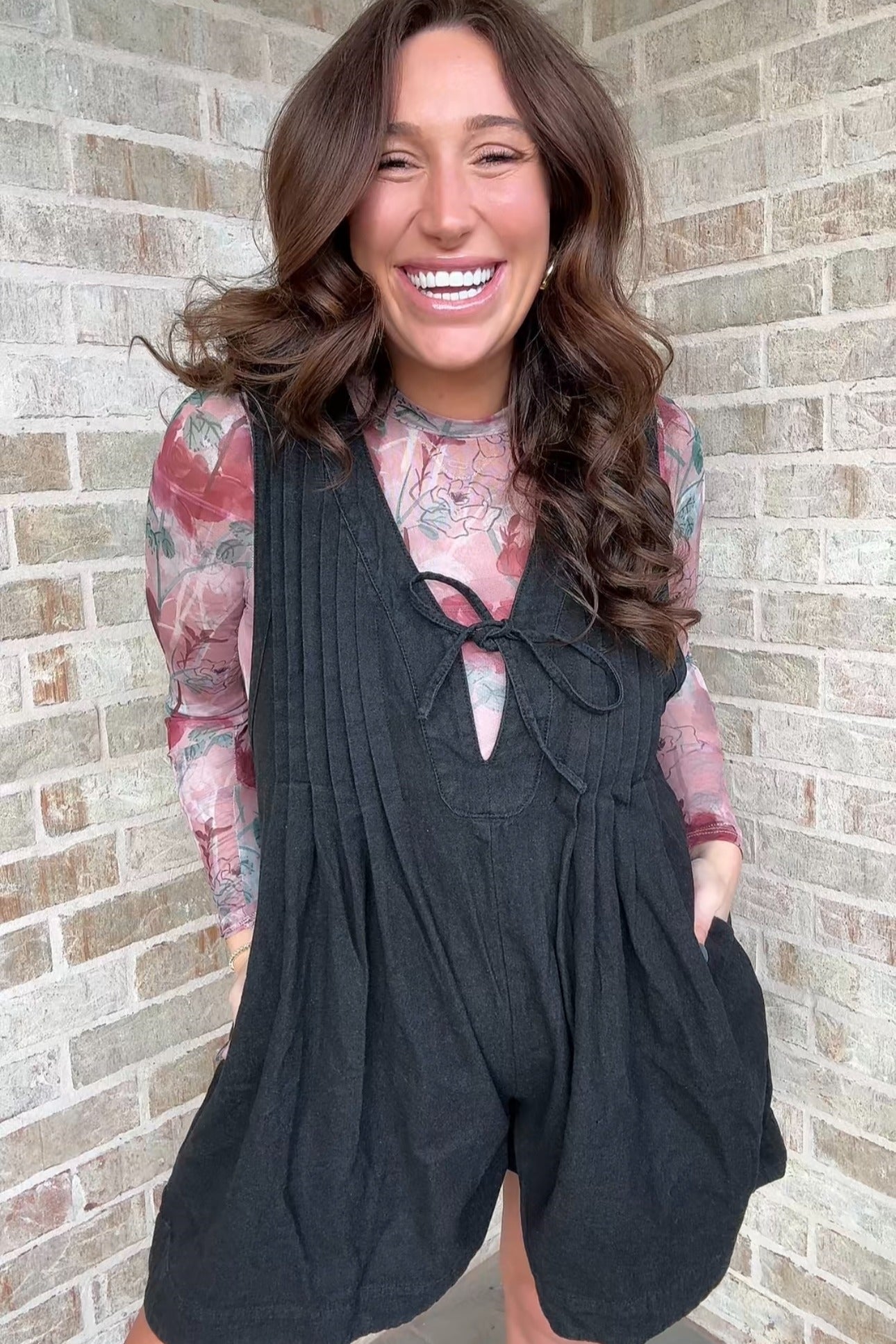 woman wearing a black maternity romper with a tie front and pockets over a floral long sleeve top