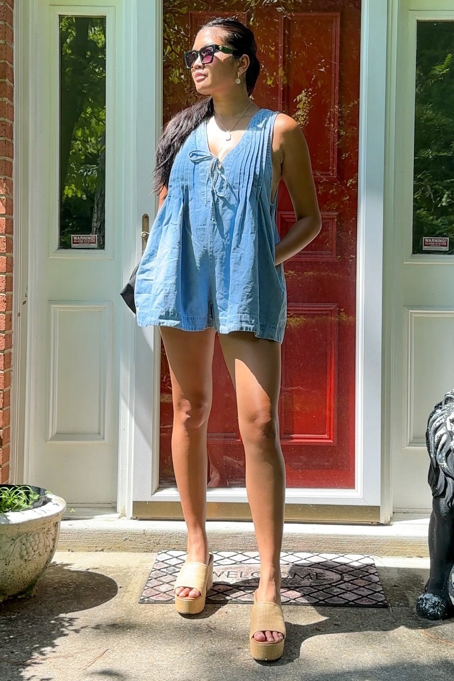 woman wearing a light blue maternity romper with a pleated front, standing in front of a red door and wearing platform sandals