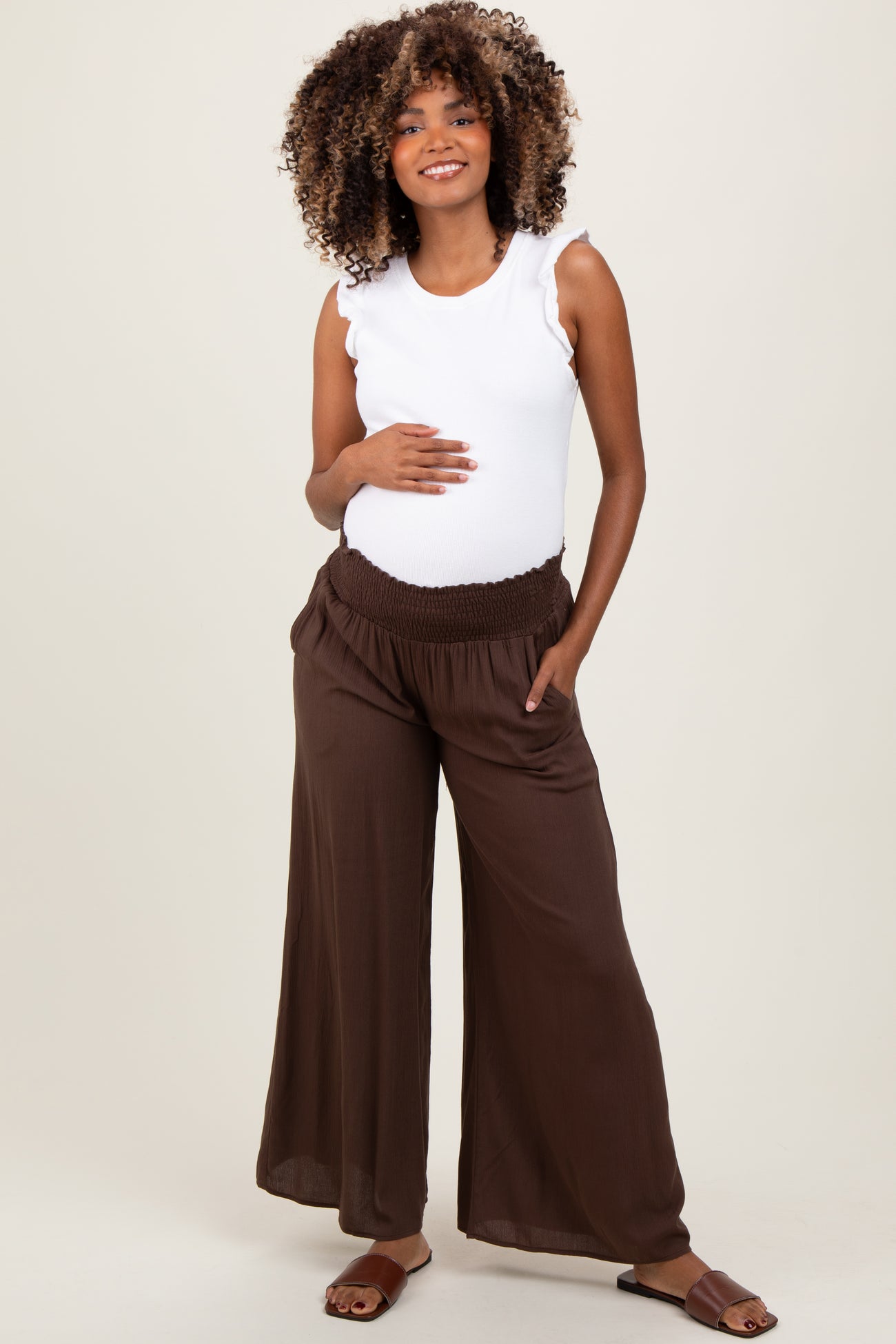 woman wearing a white sleeveless maternity top and brown wide-leg pants, gently cradling her baby bump