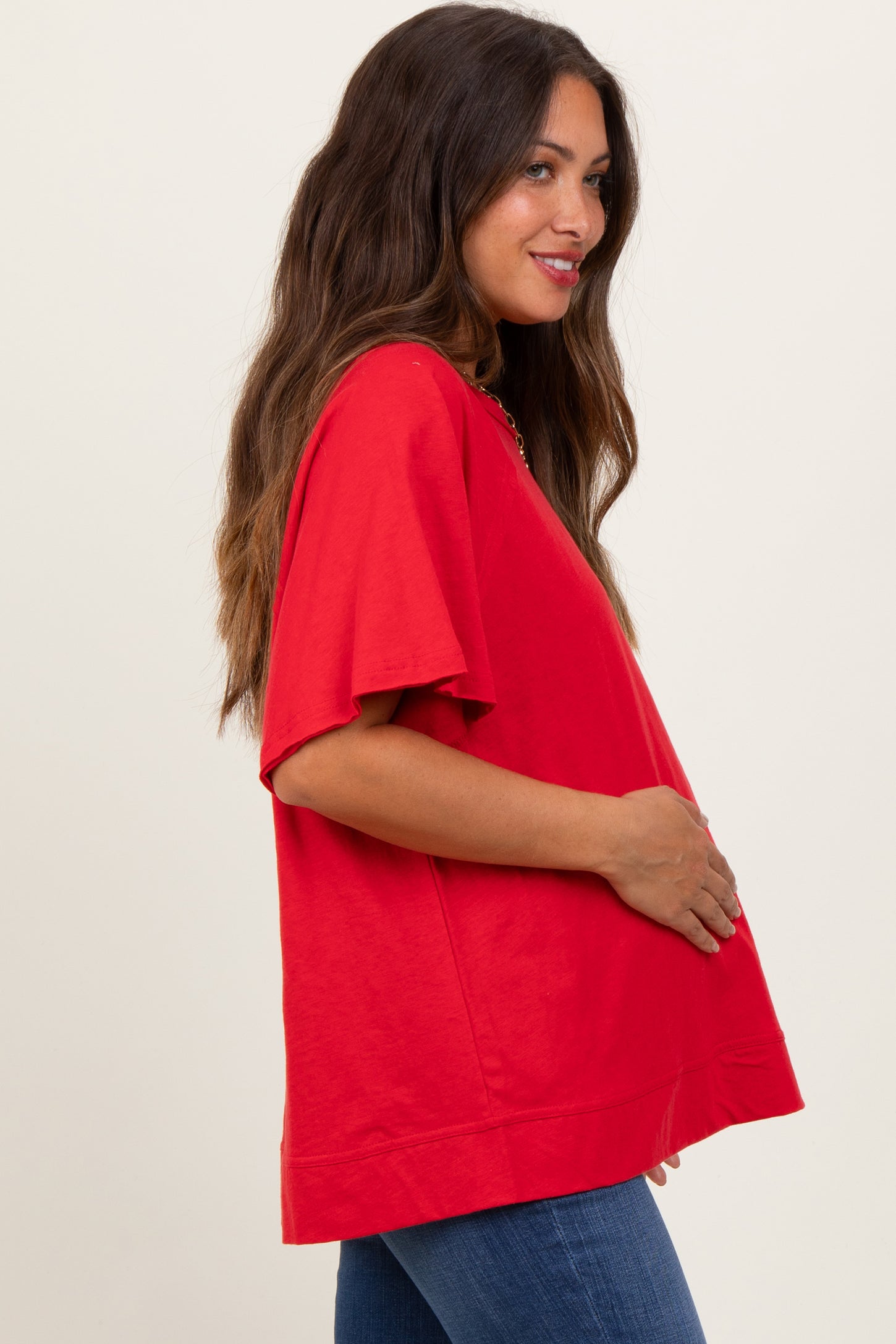 woman wearing a bright red oversized maternity top and holding her baby bump, paired with blue jeans