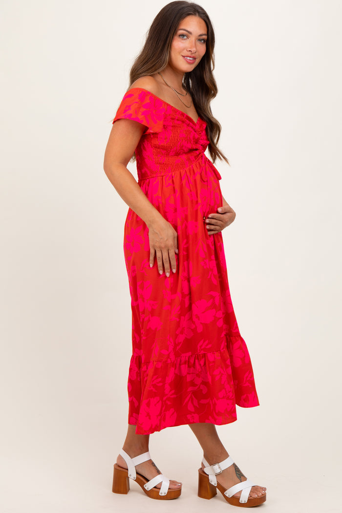 woman wearing a bright pink floral maternity dress with off-shoulder sleeves and a ruffled hem, holding her baby bump