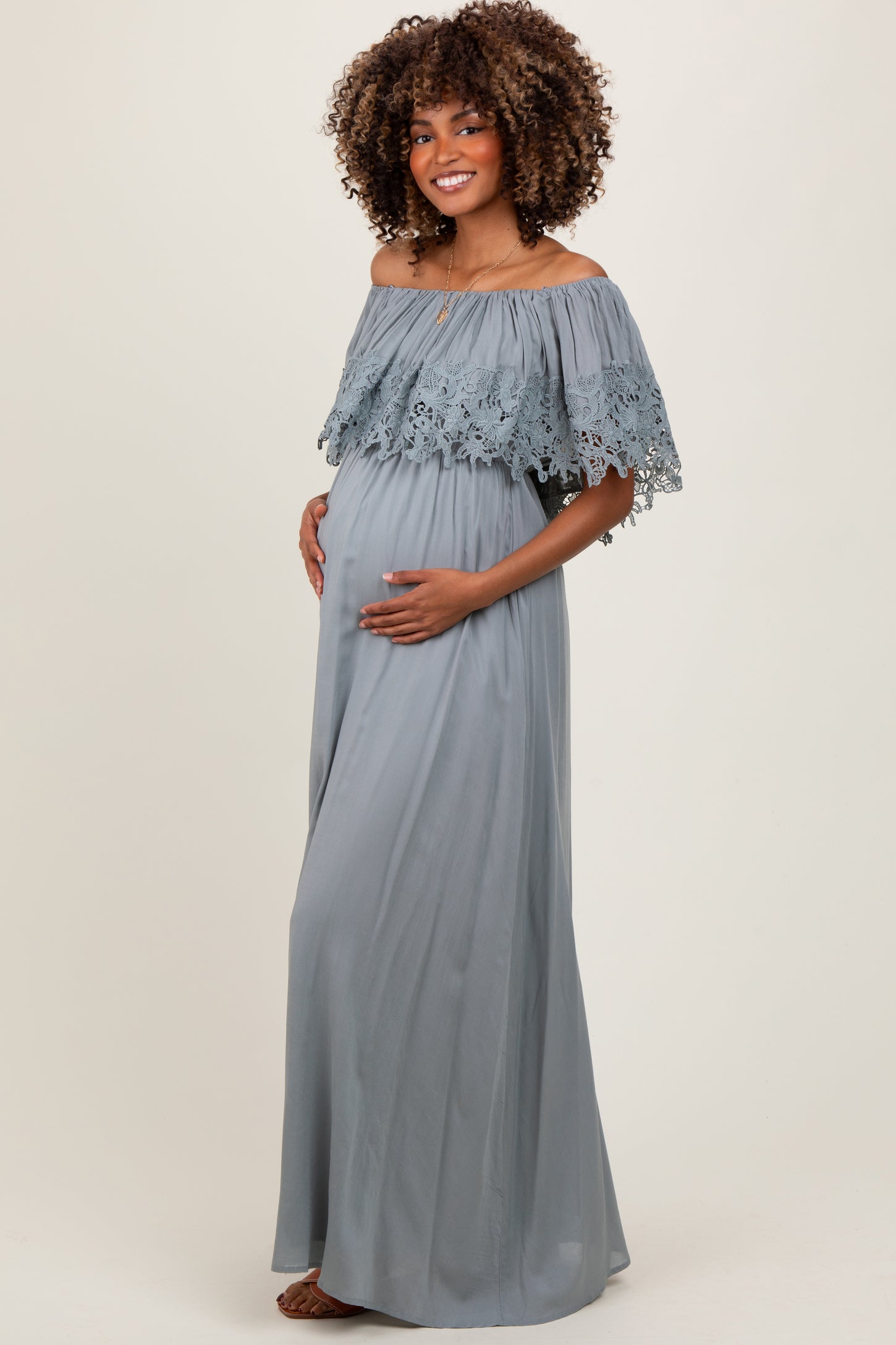 woman wearing a light gray off-the-shoulder maternity gown with lace details and holding her baby bump