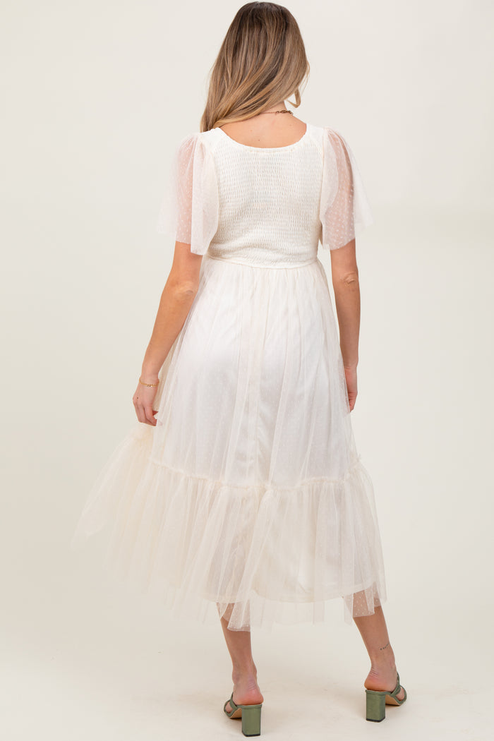 woman wearing a white maternity midi dress with sheer puff sleeves and a tiered tulle skirt, showing her baby bump