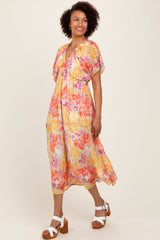 woman wearing a floral midi dress with short flutter sleeves in shades of pink, yellow, and cream, paired with white block-heeled sandals