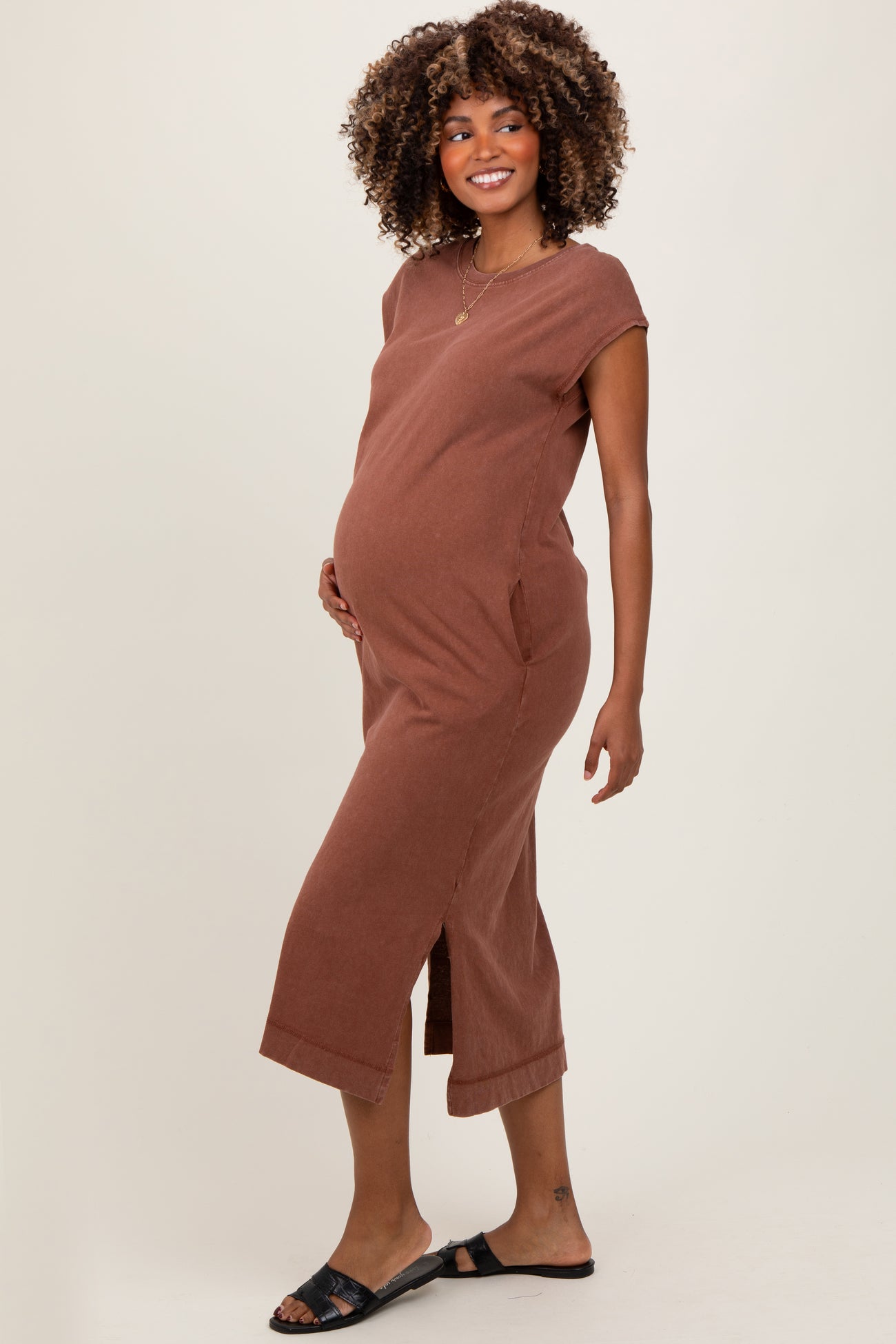 woman wearing a rust-colored maternity midi dress with short sleeves and side pockets, holding her baby bump