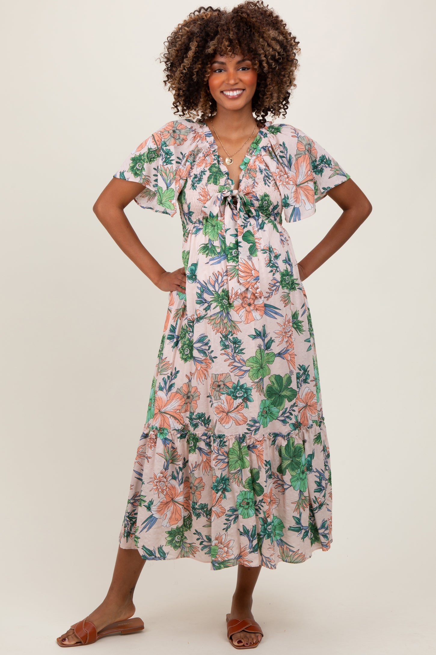 woman wearing a floral maternity dress with a tiered skirt and flutter sleeves, smiling and standing with hands on hips
