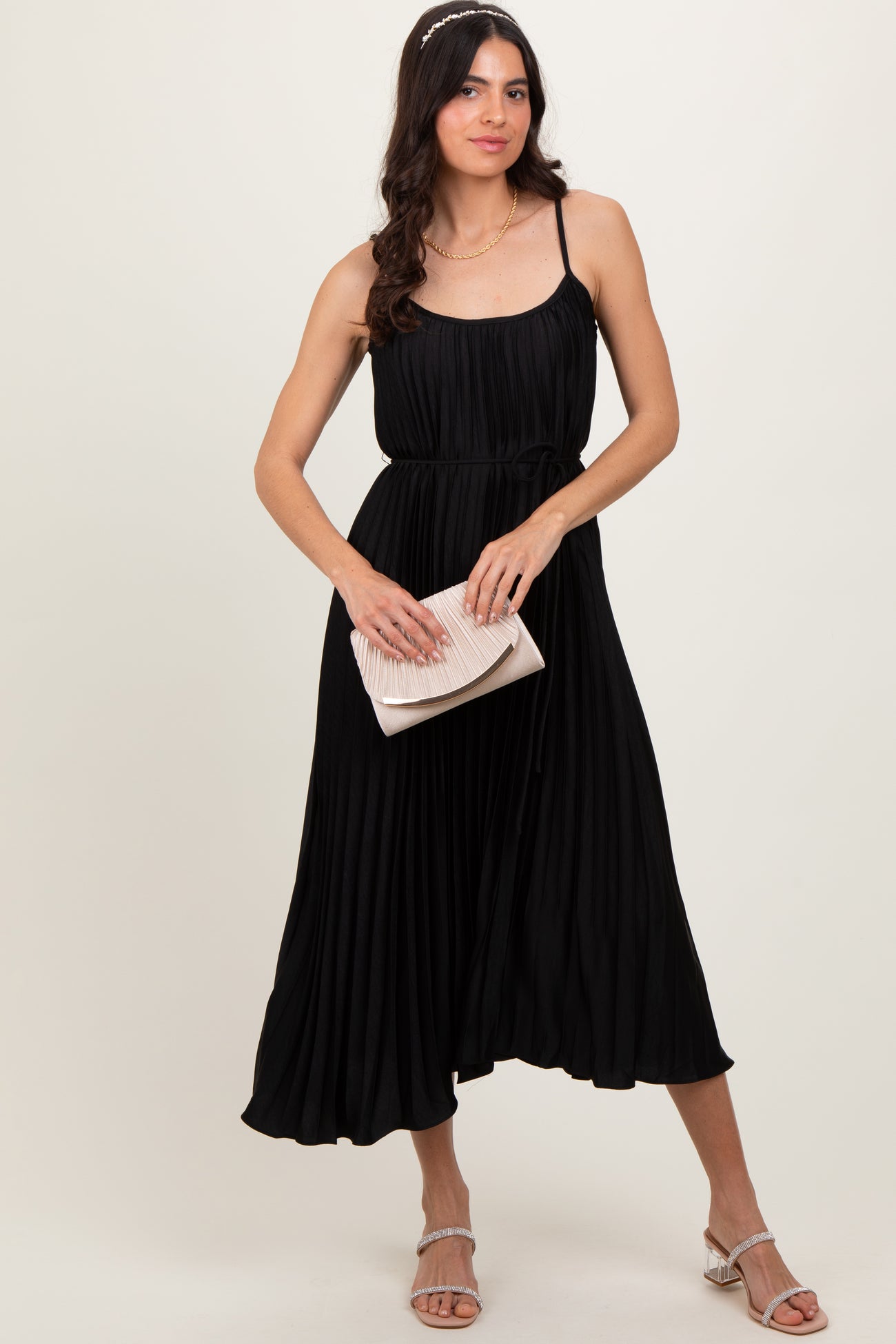woman wearing a black pleated maxi dress with spaghetti straps, holding a clutch and standing in a relaxed pose