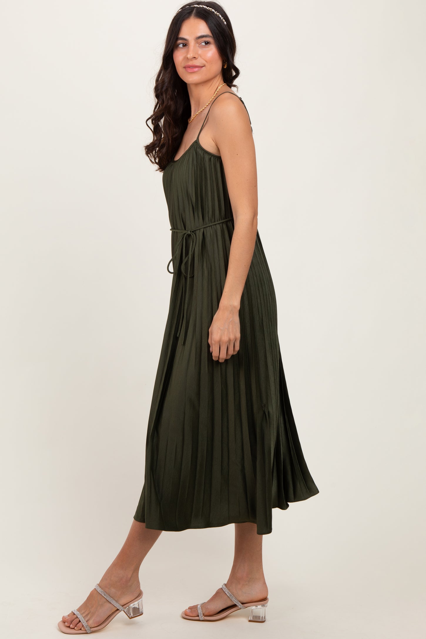 woman wearing an olive green midi dress with a tied waist and pleated skirt, paired with silver strappy sandals