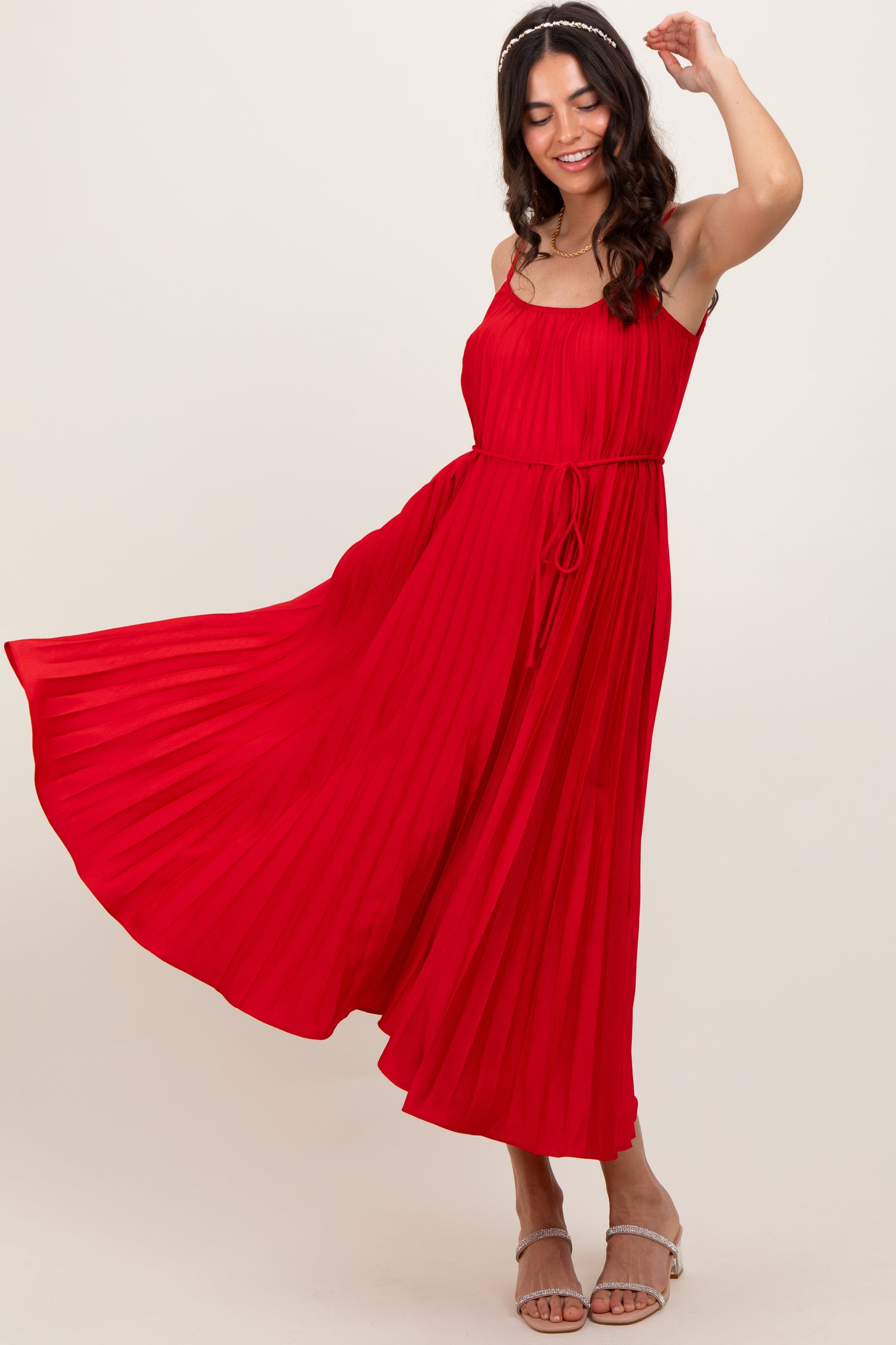 woman wearing a red pleated maxi dress with thin straps and a tie waist, smiling and twirling
