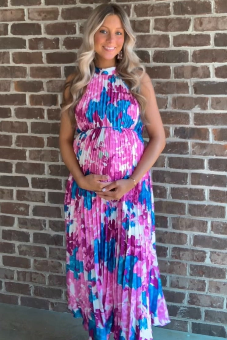 woman wearing a floral print maternity dress in pink and blue, cradling her baby bump against a brick wall
