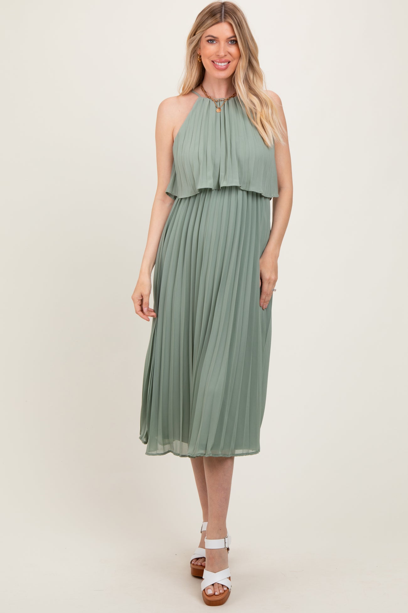 woman wearing a sage green pleated halter neck midi dress with a layered top and white heeled sandals