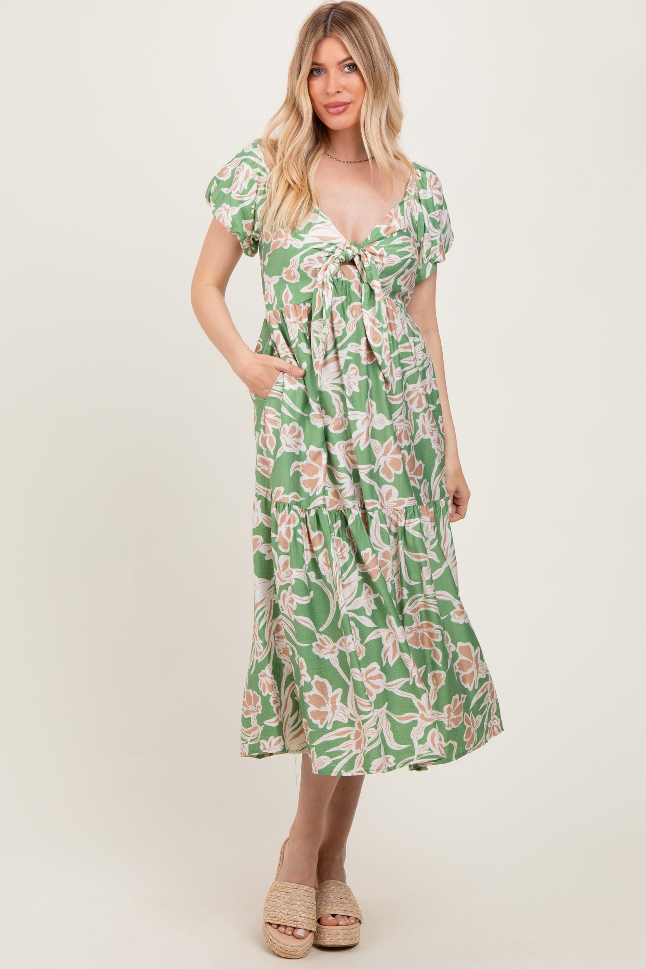 woman wearing a green floral print maxi dress with a tie front and short sleeves, standing and posing with hands in pockets