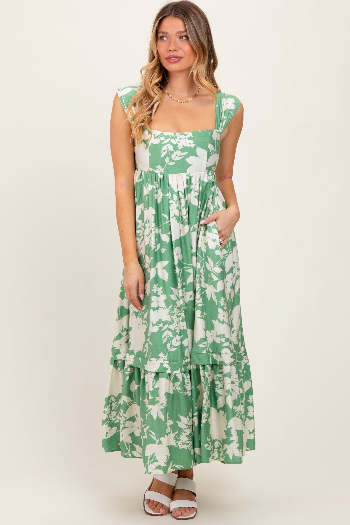 woman wearing a green floral print maxi dress with a square neckline and tiered skirt, posing with hands in pockets