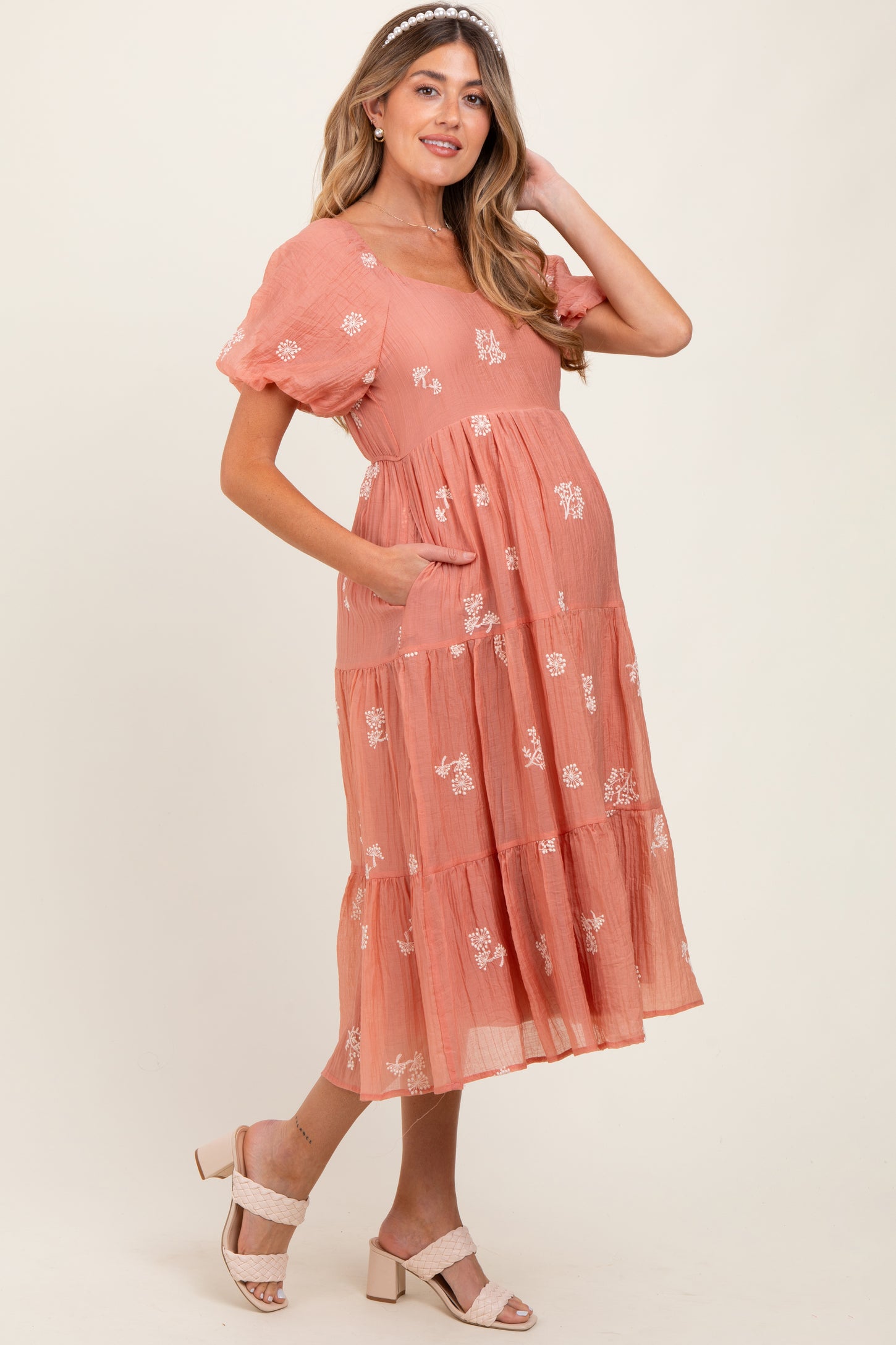 woman wearing a coral maternity dress with short sleeves, floral embroidery, and pockets, highlighting her baby bump