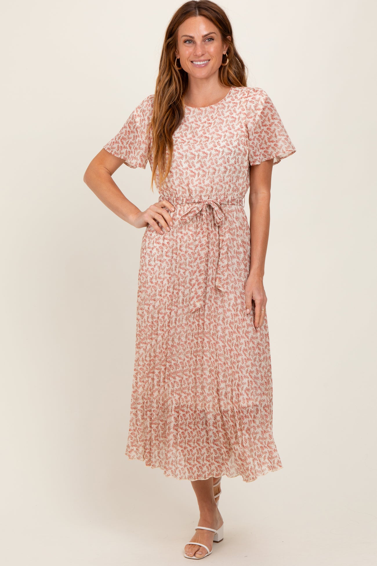 woman wearing a pink floral maxi dress with short sleeves and a tie belt, standing and smiling at the camera