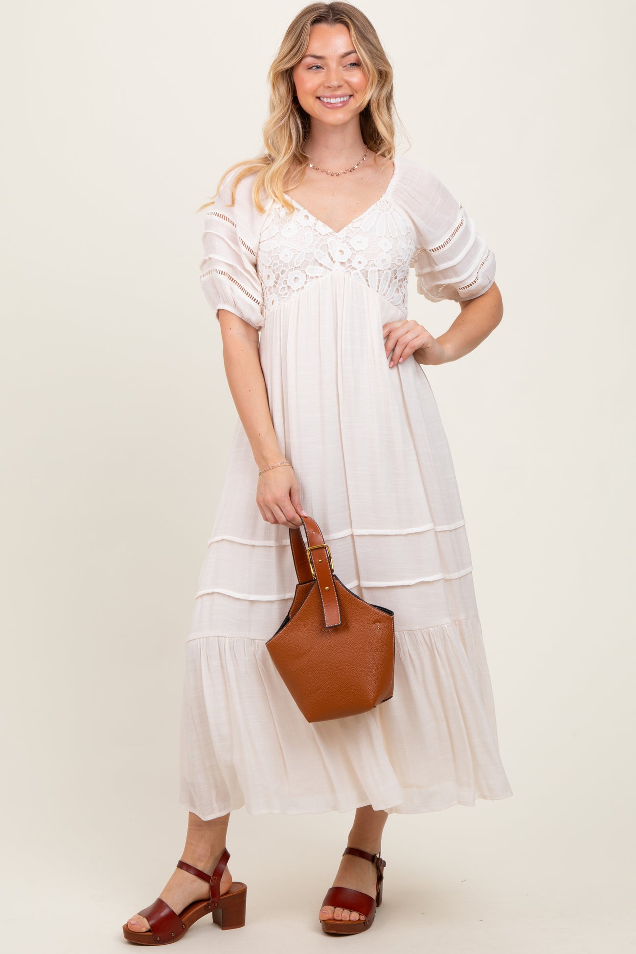 woman wearing a white maxi dress with lace detailing, holding a brown handbag and wearing brown heeled sandals