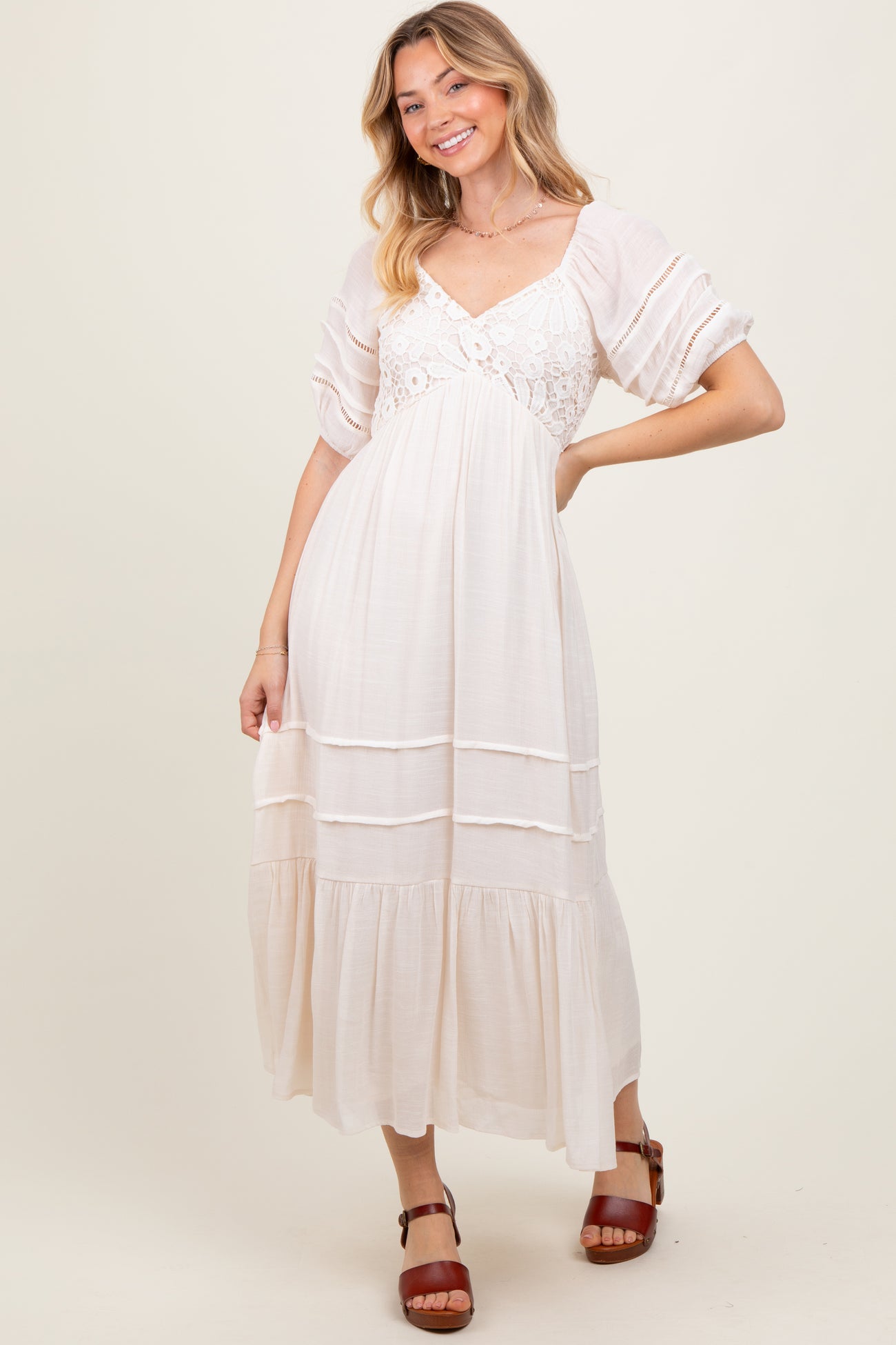 woman wearing a white maxi dress with lace details and puff sleeves, smiling and posing against a beige background
