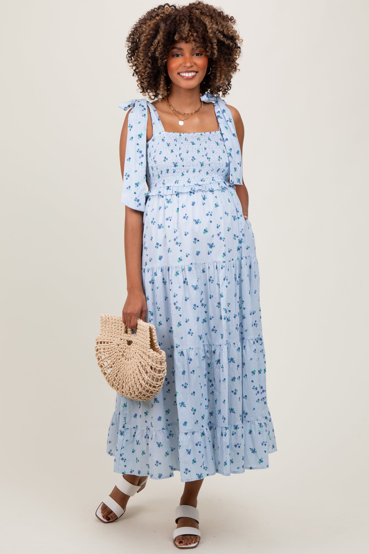 woman wearing a blue floral maternity maxi dress with a smocked bodice and tie straps, holding a woven bag