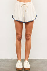 woman wearing cream-colored shorts with blue pom-pom trim and drawstring waist, paired with white sneakers