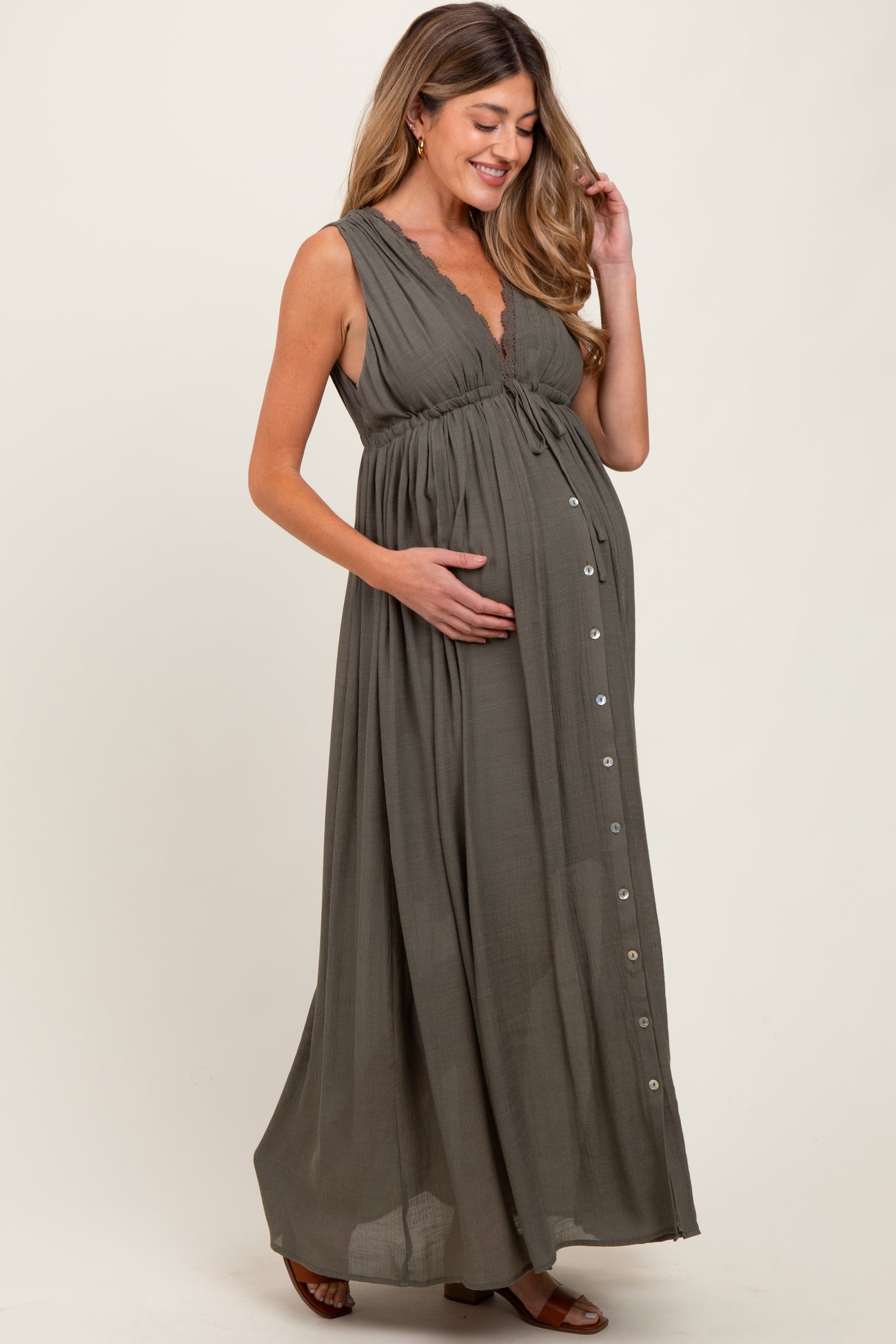 woman wearing an olive green sleeveless maternity maxi dress with a button detail and crinkle fabric, holding her baby bump