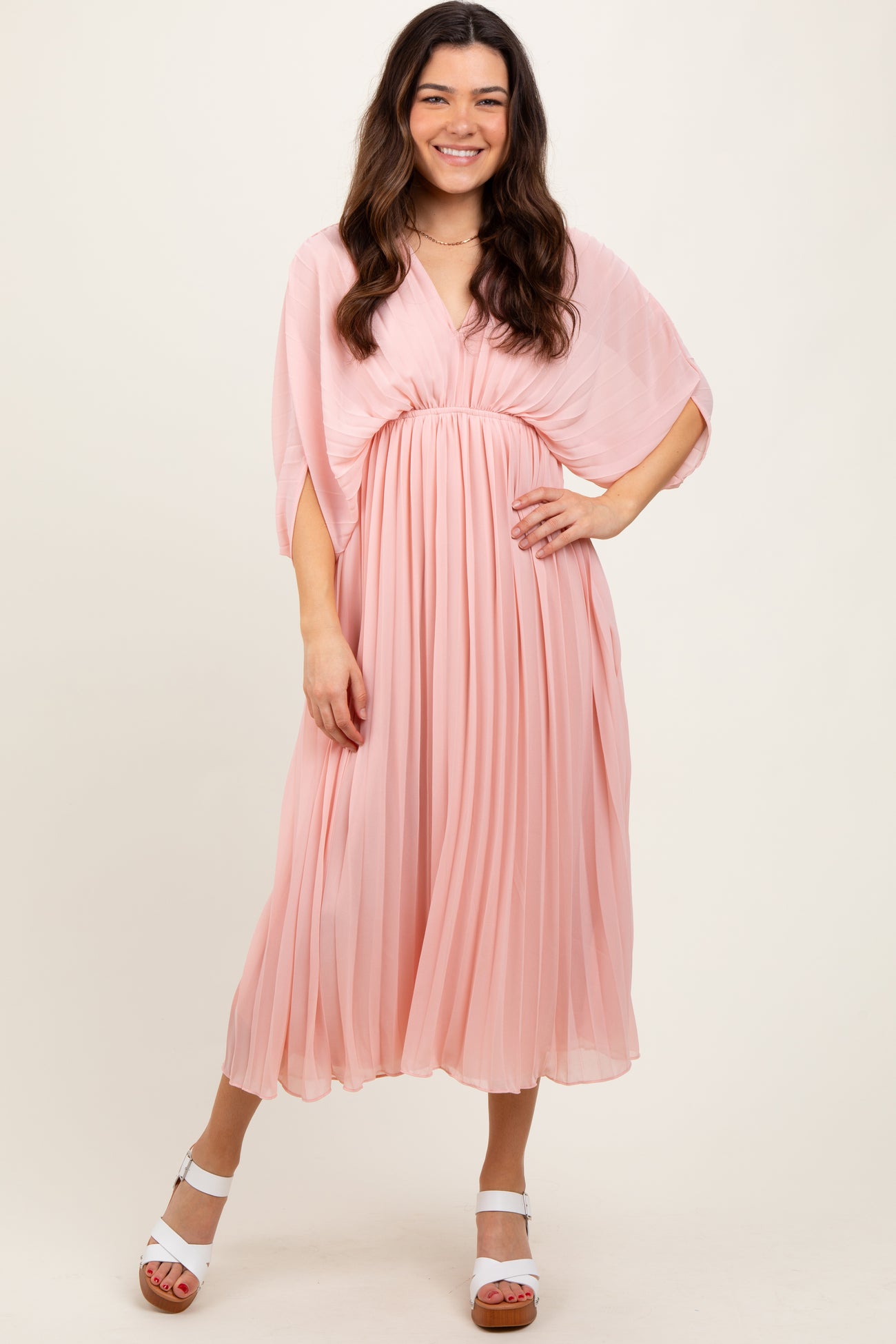 woman wearing a light pink pleated midi dress with a flowy cape sleeve and a cinched waist