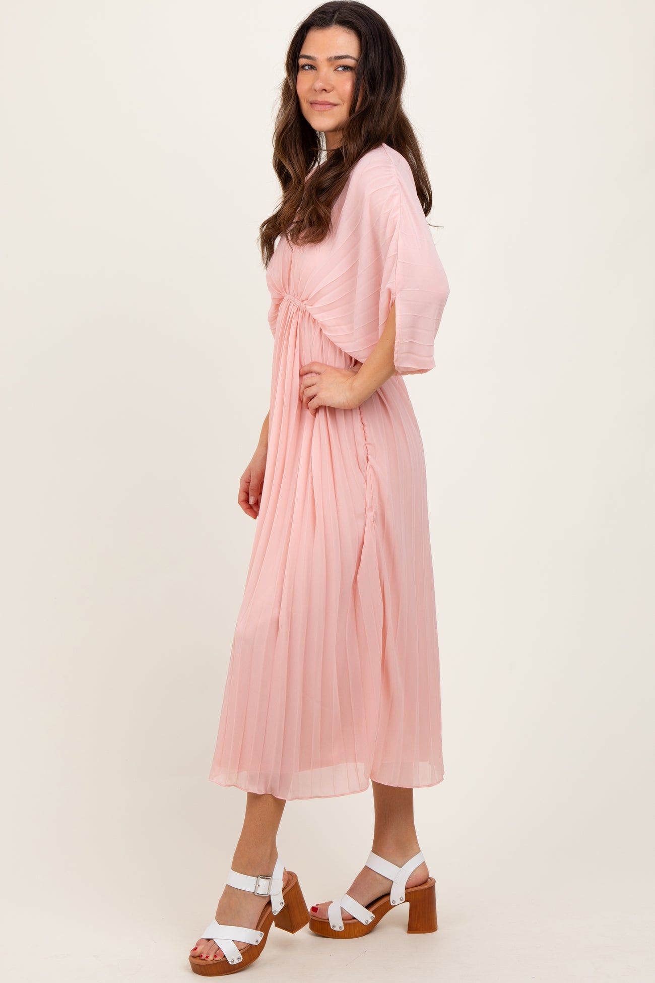 woman wearing a light pink pleated midi dress with short sleeves and a tied waist, paired with white block-heeled sandals
