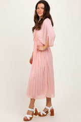 woman wearing a light pink pleated midi dress with short sleeves and a tied waist, paired with white block-heeled sandals