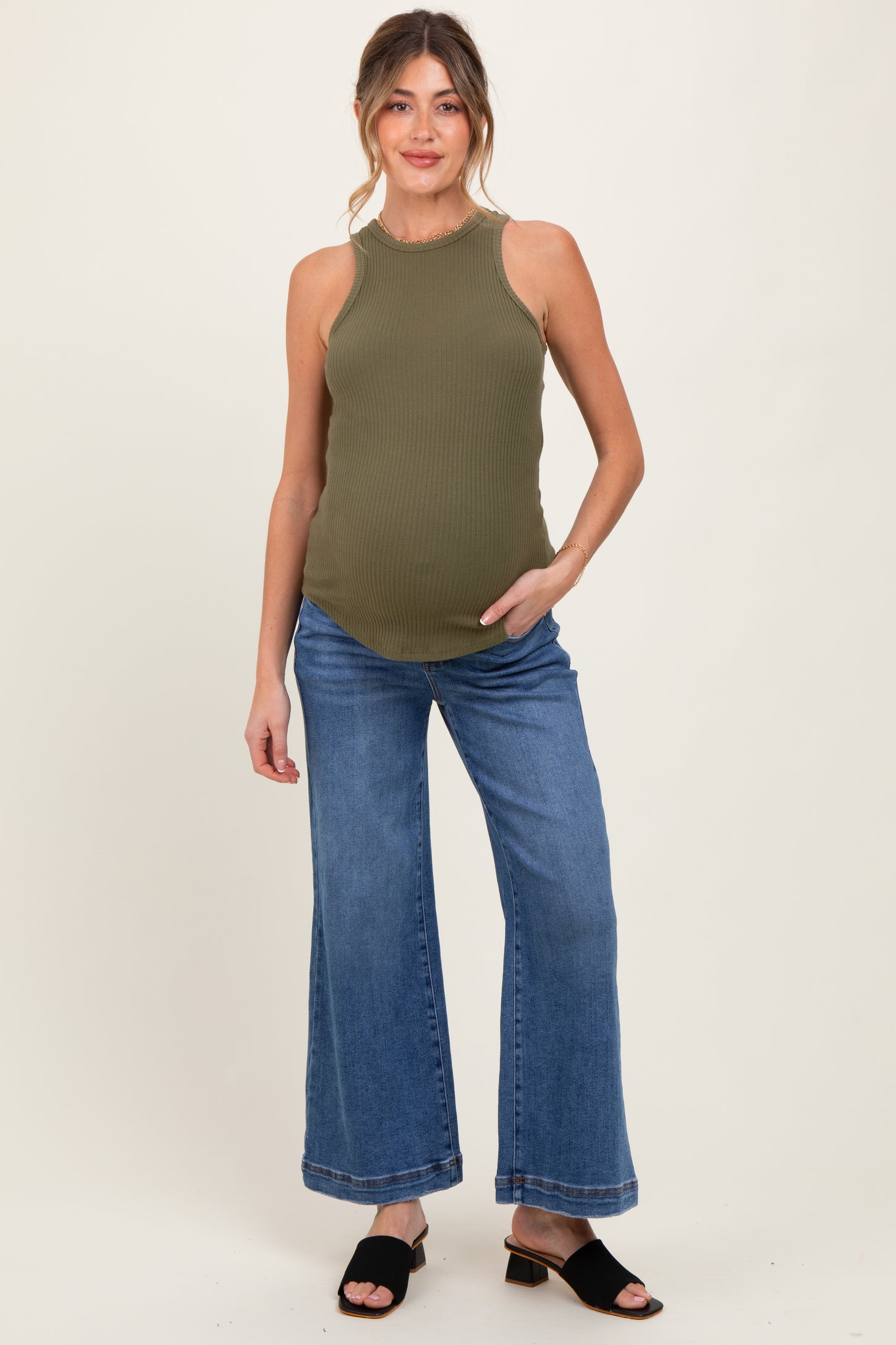 woman wearing a dark green ribbed maternity tank top and light blue wide-leg jeans, holding her baby bump