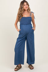 woman wearing a blue striped maternity jumpsuit with a smocked bodice and side pockets, featuring adjustable straps and a baby bump visible