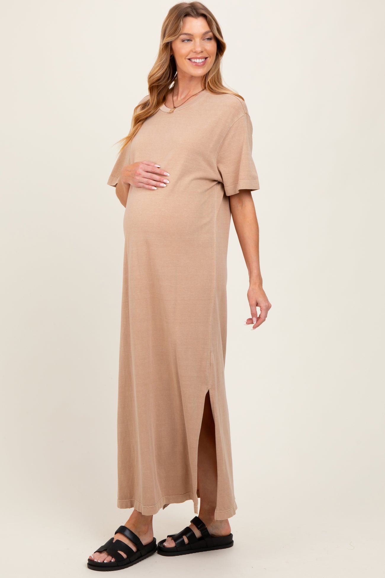 woman wearing a beige short sleeve maternity maxi dress with side slits and holding her baby bump