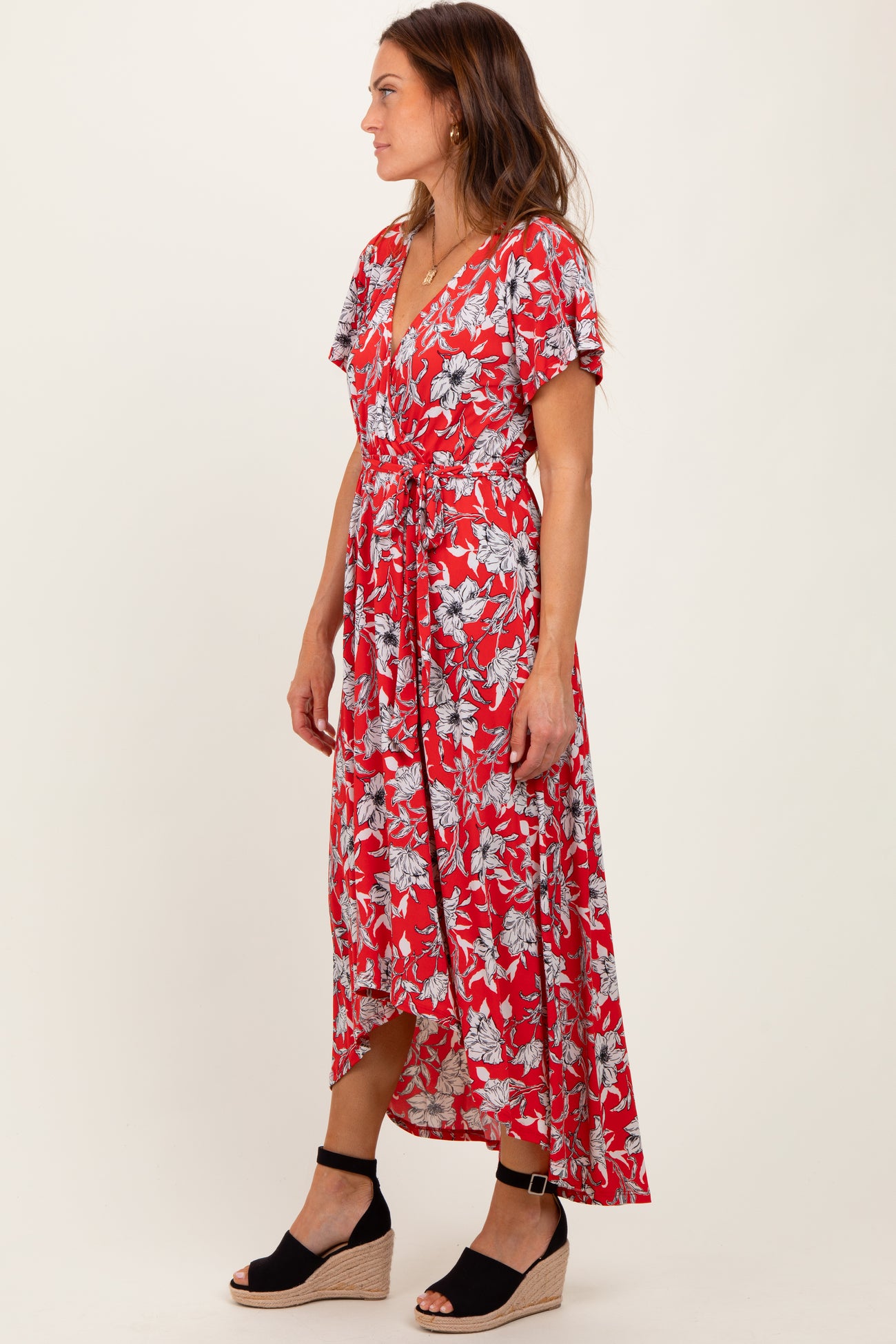 woman wearing a red floral wrap maxi dress with flutter sleeves and a tied waist, standing in profile on a plain background