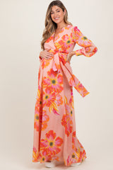 woman wearing a floral pink maternity maxi dress with long sleeves and a tie waist, holding her baby bump
