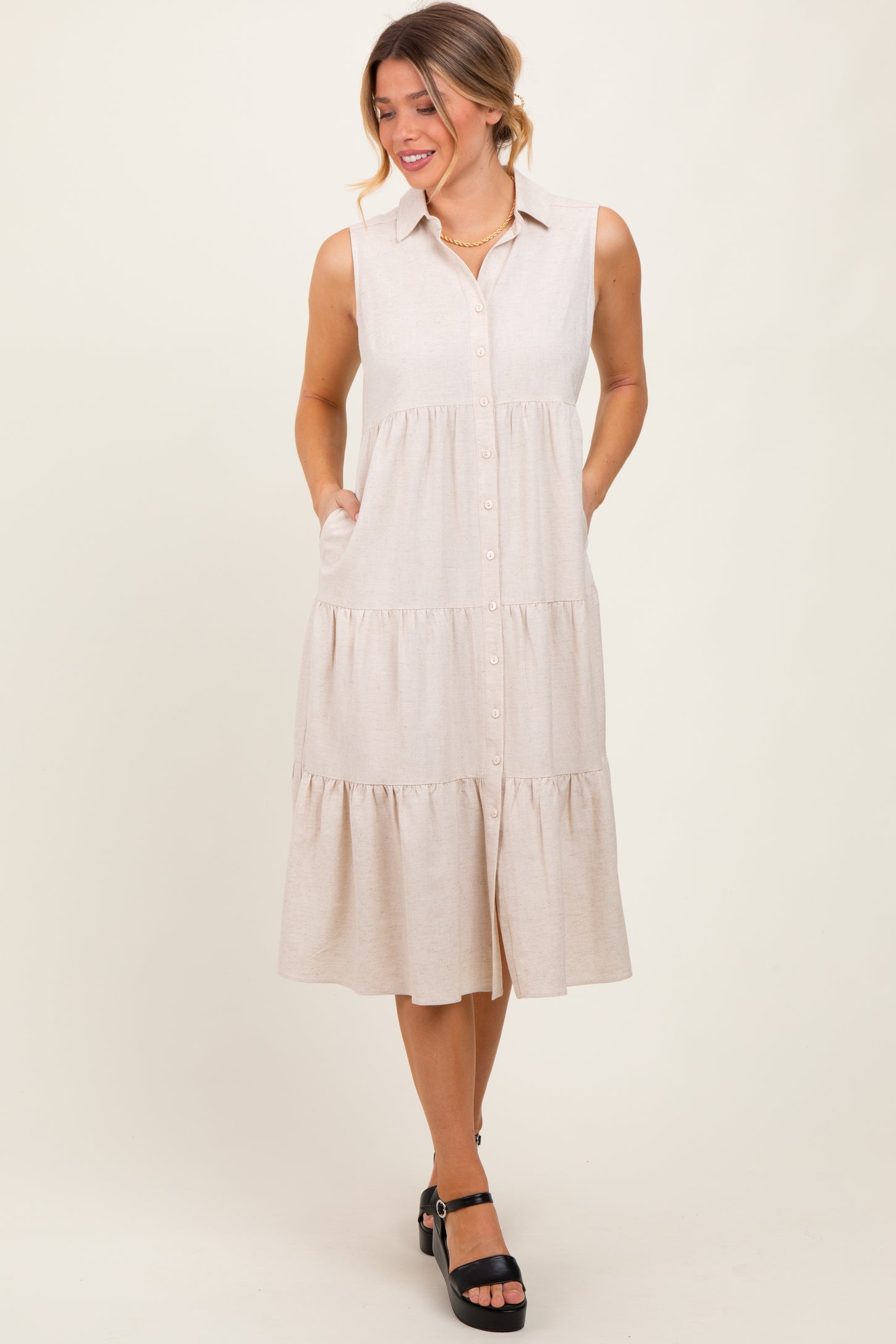woman wearing a beige sleeveless button-up tiered midi dress with side pockets and black platform heels