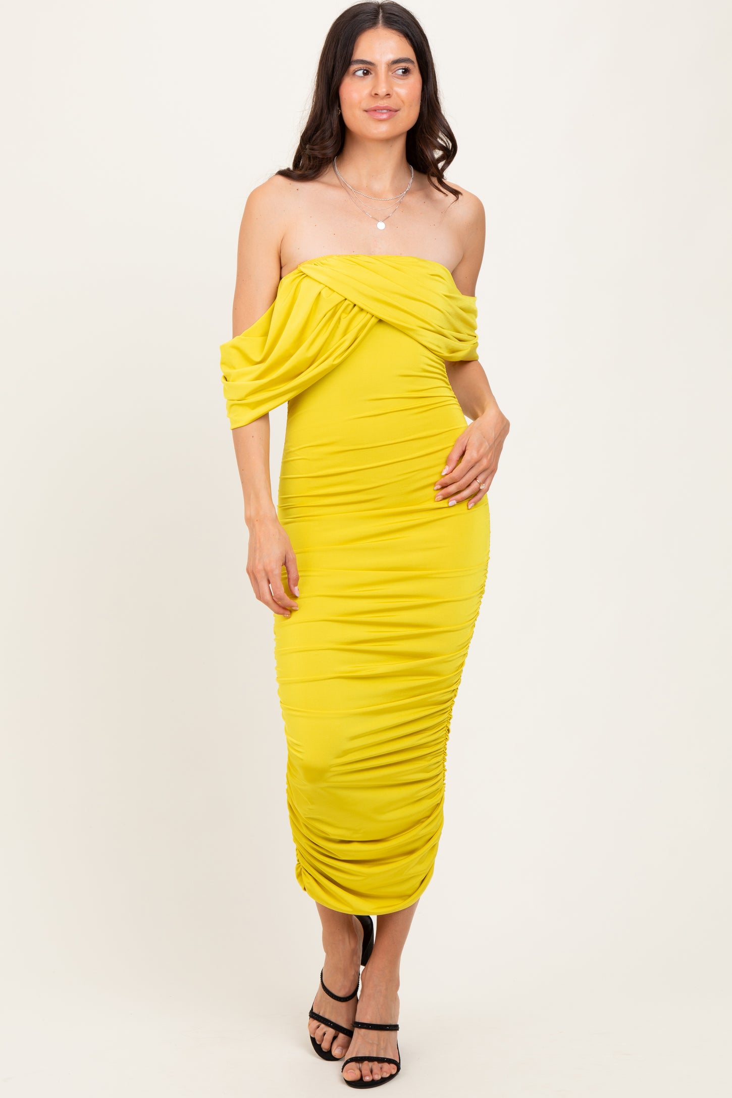 woman wearing a yellow off-shoulder ruched midi dress with a form-fitting silhouette and strappy black heels