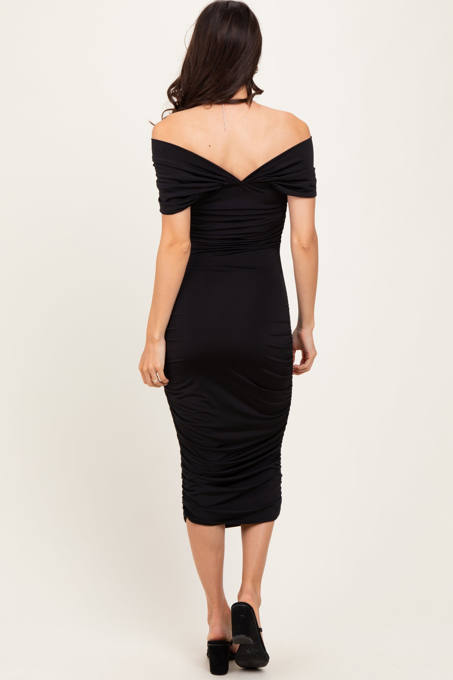 woman wearing a black off-the-shoulder ruched midi dress, showing the back view and fitted silhouette