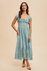 woman wearing a teal floral maxi dress with ruffle sleeves and pockets, standing against a neutral background