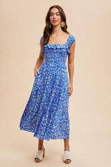 woman wearing a floral print blue midi dress with ruffle straps and pockets, standing against a neutral background