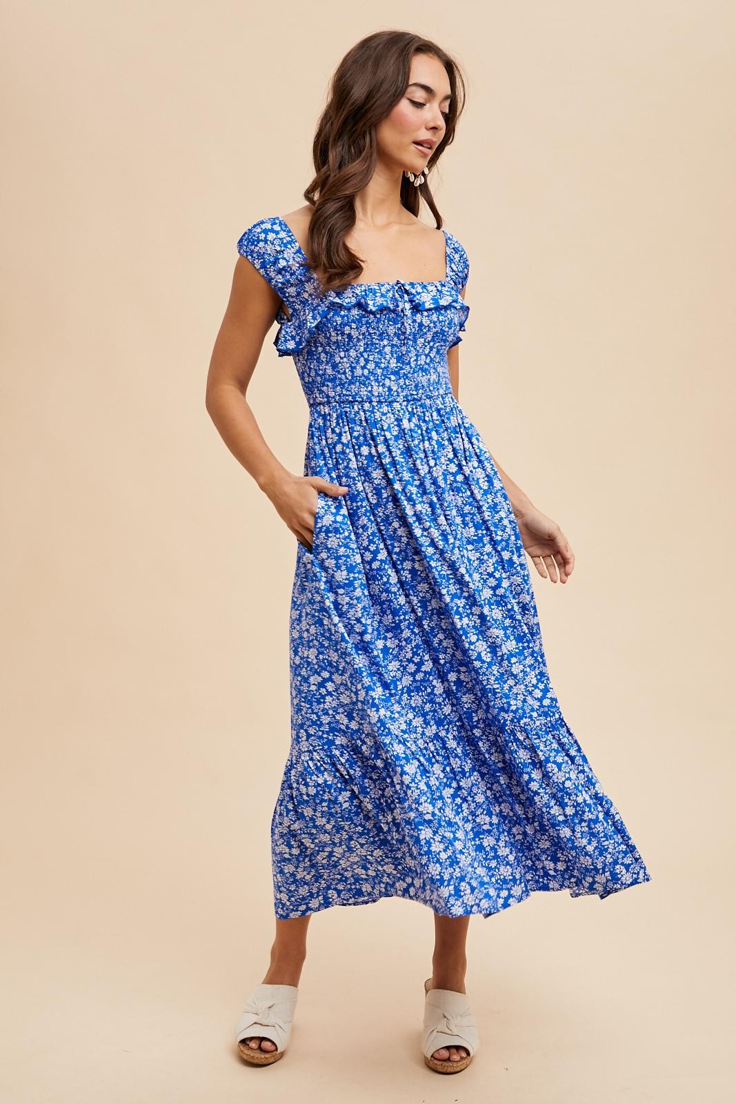 woman wearing a blue floral midi dress with ruffle sleeves and pockets, standing against a neutral background