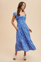 woman wearing a blue floral midi dress with ruffle sleeves and pockets, standing against a neutral background