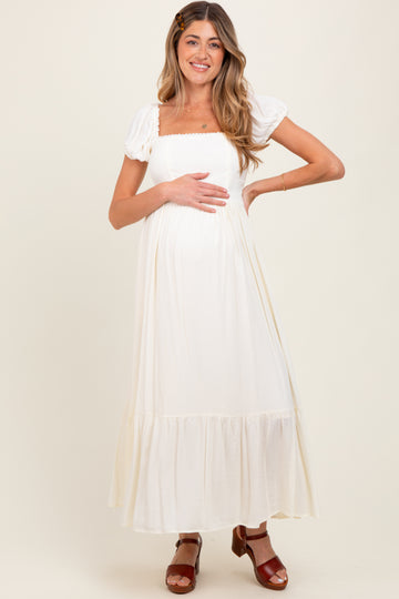 woman wearing a white maternity maxi dress with puff sleeves and a gathered waist, cradling her baby bump